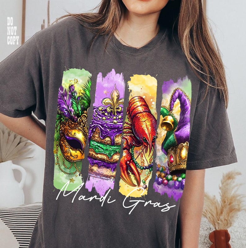 May include: Dark gray t-shirt featuring a colorful Mardi Gras design. The design includes a mask, cake, crawfish, and jester hat, with the words "Mardi Gras" in white script. The shirt is a comfortable, casual style.