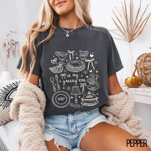 May include: Dark grey t-shirt with a whimsical graphic of kitchenware and the text "in my + granny era." The design includes teacups, plates, and a duck. A vintage-inspired tee, perfect for a cosy look.