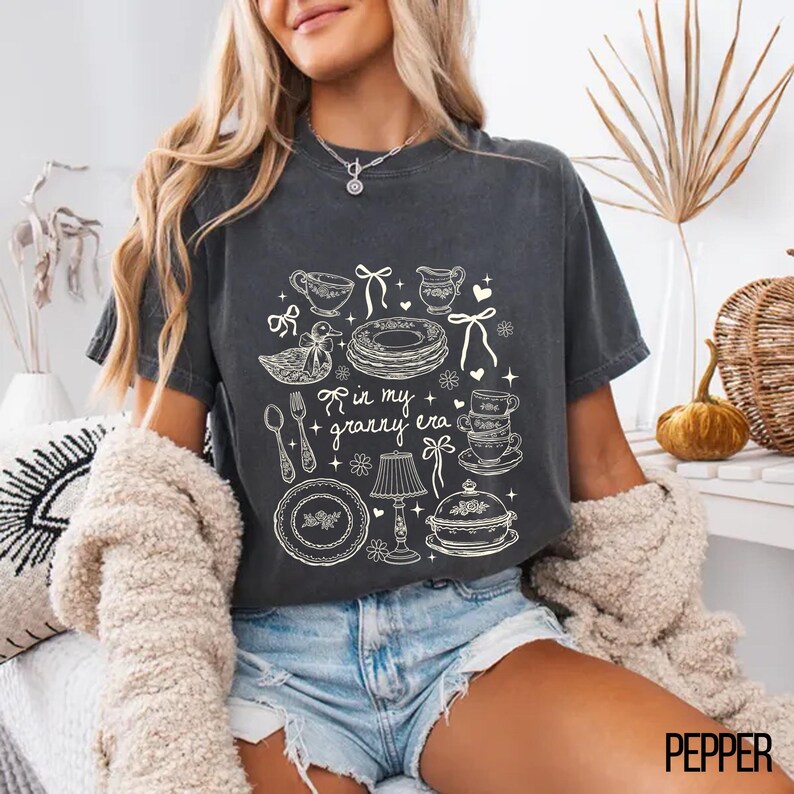 May include: Dark grey t-shirt with a whimsical graphic of kitchenware and the text "in my + granny era." The design includes teacups, plates, and a duck. A vintage-inspired tee, perfect for a cosy look.