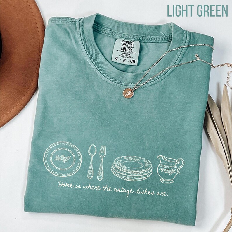 May include: Light green Comfort Colors t-shirt with a white graphic of vintage dishes and the phrase "Home is where the vintage dishes are." A gold necklace with a pendant rests on the shirt.