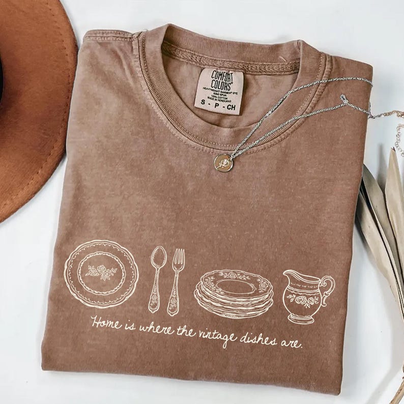 May include: A brown Comfort Colors t-shirt with a white graphic of vintage dishes and the phrase "Home is where the vintage dishes are." A silver necklace with a pendant is draped across the shirt.