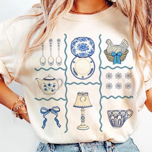 Pode incluir: Camisola creme com uma grelha de ilustra&ccedil;&otilde;es azuis e brancas. O design inclui colheres, pratos, uma chaleira, uma galinha numa cesta, um la&ccedil;o, uma l&acirc;mpada, uma ch&aacute;vena de ch&aacute; e detalhes florais. O tema geral &eacute; utens&iacute;lios de cozinha vintage.