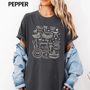 May include: Dark grey t-shirt featuring a white graphic of kitchen items such as teacups and plates, with the text "in my granny era". The shirt has short sleeves and a loose fit. The word "pepper" is printed at the top.