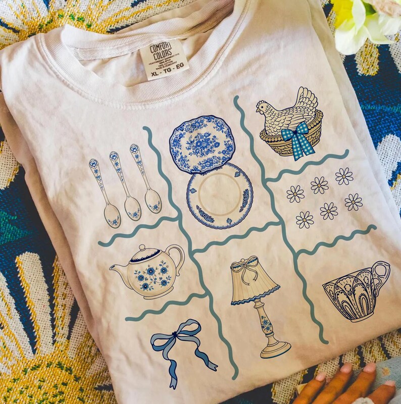 Pode incluir: Camiseta creme XL Comfort Colors com um design de grade com ilustra&ccedil;&otilde;es azuis e brancas. O design inclui pratos, colheres, uma chaleira, uma l&acirc;mpada, uma x&iacute;cara, uma galinha em uma cesta, flores e um la&ccedil;o.