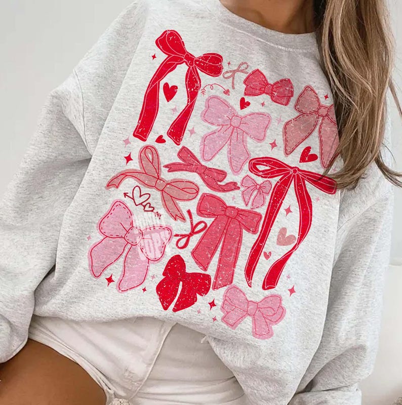 May include: A light gray sweatshirt featuring a pattern of red and pink bows, hearts, and stars. The bows vary in size and style, with some appearing stitched. The design is spread across the front of the sweatshirt, creating a playful and feminine aesthetic.