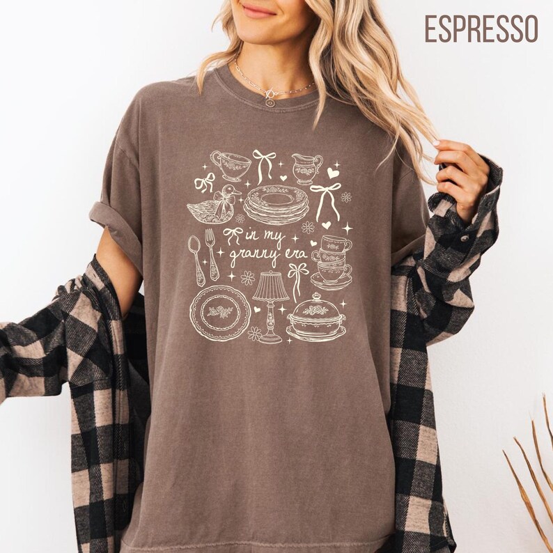 May include: Espresso-coloured t-shirt with a white graphic design featuring kitchen and dining elements. The design includes a duck, plates, cups, and the text "in my granny era". The shirt has short sleeves and a relaxed fit.
