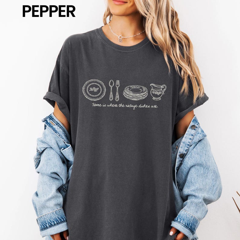 May include: Dark gray t-shirt with a white graphic of vintage dishes, including a plate, silverware, and a pitcher. The text "Home is where the vintage dishes are" is below the graphic. The shirt has rolled-up sleeves and is worn with a denim jacket.