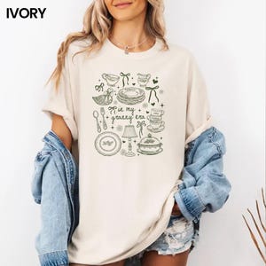 May include: Cream-coloured t-shirt featuring a green illustration of kitchen items, such as plates and cups, and a duck. The text "in my + granny era" is printed on the front. The shirt has short sleeves and a relaxed fit.