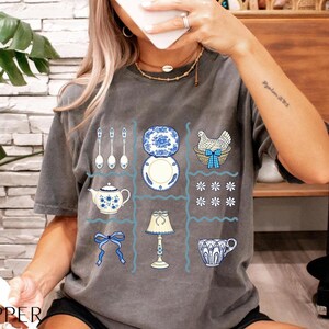 Pode incluir: Camiseta cinza com um design de grade apresentando ilustra&ccedil;&otilde;es azuis e brancas de utens&iacute;lios de cozinha e itens decorativos. O design inclui colheres, pratos, uma chaleira, uma l&acirc;mpada, uma x&iacute;cara, um la&ccedil;o e uma galinha em uma cesta.