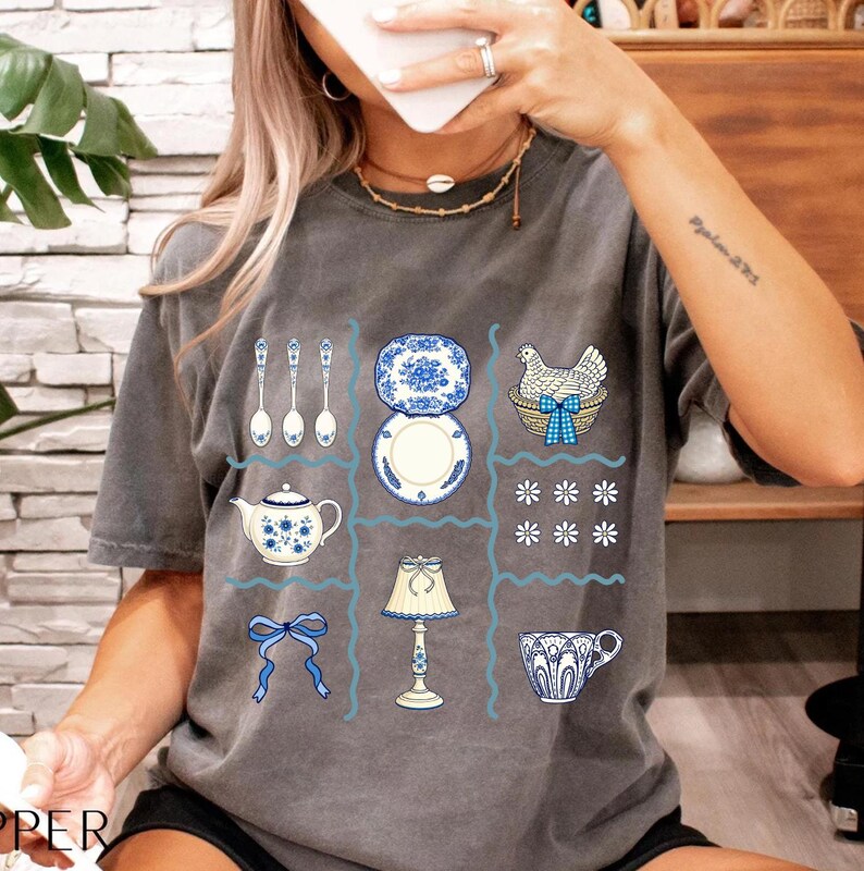 Pode incluir: Camiseta cinza com um design de grade apresentando ilustra&ccedil;&otilde;es azuis e brancas de utens&iacute;lios de cozinha e itens decorativos. O design inclui colheres, pratos, uma chaleira, uma l&acirc;mpada, uma x&iacute;cara, um la&ccedil;o e uma galinha em uma cesta.