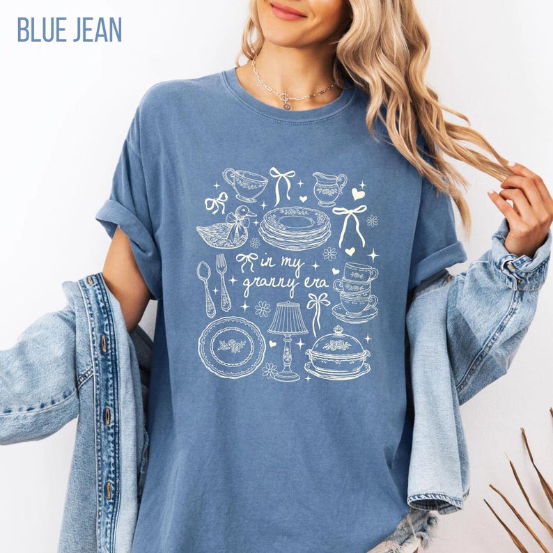 May include: A blue jean coloured t-shirt with a white graphic design. The design includes plates, teacups, and a duck, with the text "in my + granny era". The t-shirt is a comfortable, casual style.