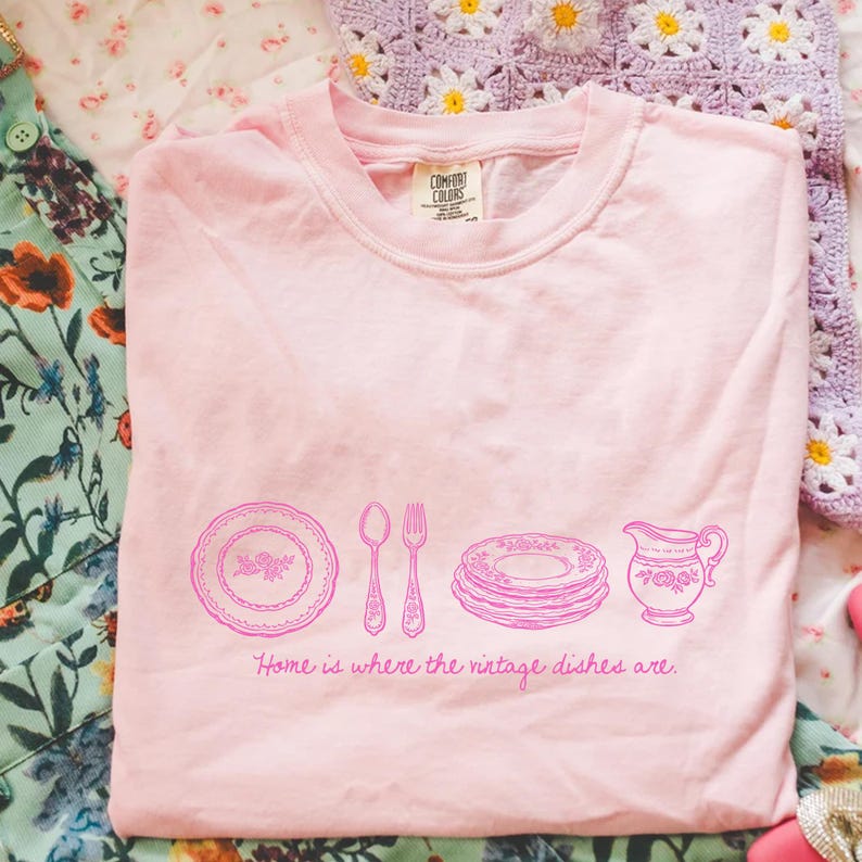 May include: A light pink t-shirt with a graphic of vintage dishes and cutlery in pink. The text "Home is where the vintage dishes are" is printed below the graphic. The shirt is made of a soft material.