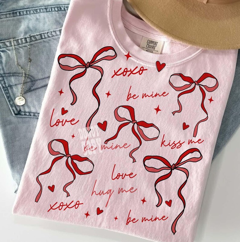 May include: A light pink t-shirt featuring red bows, hearts, and text. The text includes phrases like "xoxo," "be mine," "love," and "kiss me." The shirt is made of a soft material and is suitable for casual wear.