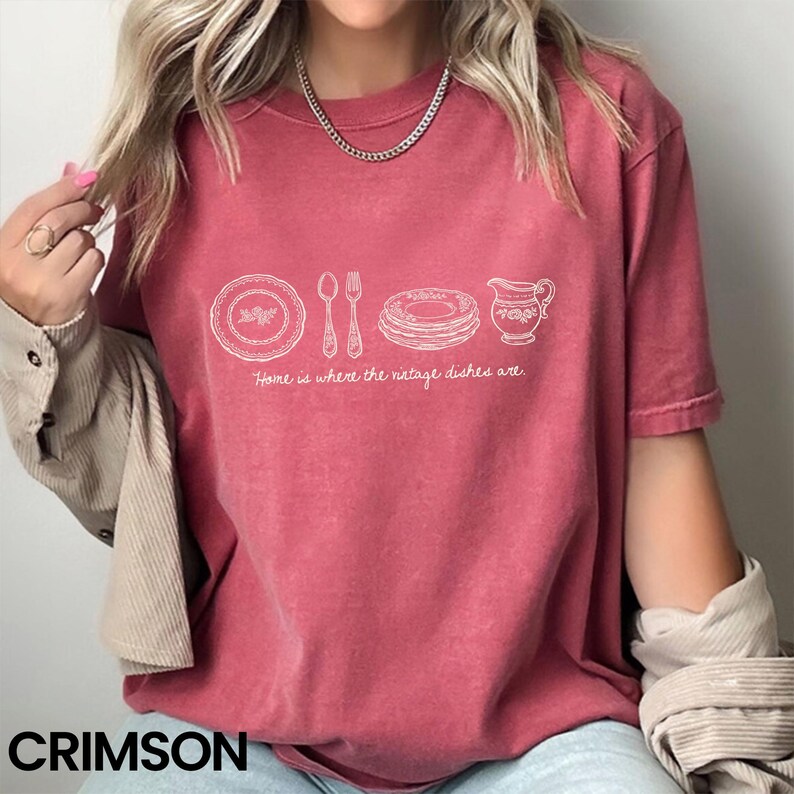May include: Crimson-colored t-shirt featuring a white line drawing of vintage dishes, including a plate, spoon, fork, stacked plates, and a pitcher. The text "Home is where the vintage dishes are" is below the drawing. The word "CRIMSON" is at the bottom.