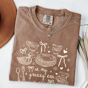 May include: A brown Comfort Colors t-shirt with a white graphic design featuring teacups, plates, a duck, and the phrase "in my granny era." A silver necklace with a pendant is draped across the shirt.