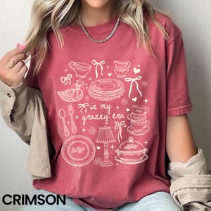 May include: Crimson t-shirt with a white graphic of kitchen items and the text "in my + granny era." The design includes teacups, plates, a duck, and a lamp. The word "CRIMSON" is printed at the bottom of the shirt.