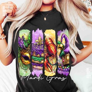 May include: Black t-shirt featuring a colorful Mardi Gras design. The design includes a gold mask with purple and green feathers, a slice of cake with a fleur-de-lis, a red crawfish, and a purple jester hat. The words "Mardi Gras" are written in white.