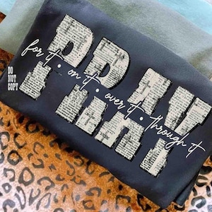 May include: Dark navy blue t-shirt with the words "PRAY OVER IT THROUGH IT" in a newspaper print design. The phrase "for it. on it." is also written on the shirt in a cursive font. The words "DO NOT COPY" are also on the shirt.