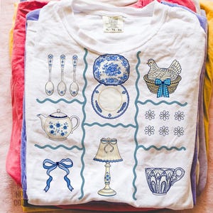 Pode incluir: Camiseta branca com um design floral azul e branco. O design inclui colheres, pratos, uma chaleira, uma galinha numa cesta, flores, uma l&acirc;mpada, um la&ccedil;o e uma ch&aacute;vena de ch&aacute;. A camisa tem uma etiqueta que diz "XL - TG - EG".