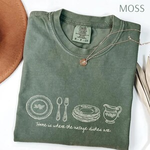 May include: Moss green t-shirt with a crew neck and short sleeves. The shirt features a white graphic of vintage dishes, a spoon, a fork, and a creamer. The text "Home is where the vintage dishes are" is printed below the graphic. A gold necklace is draped across the shirt.
