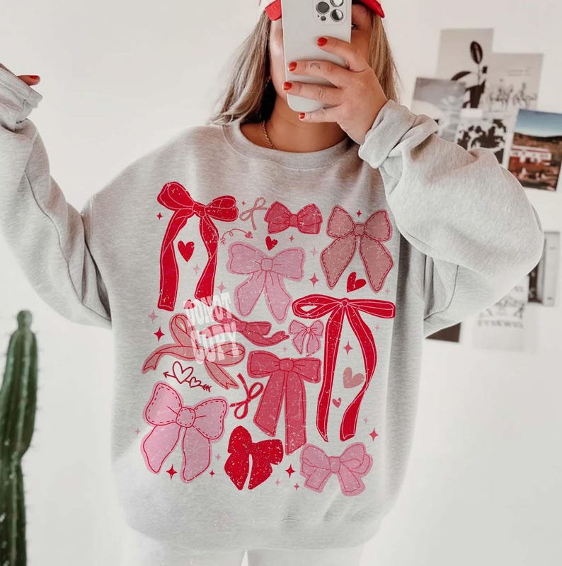 May include: A light gray sweatshirt featuring a collection of pink and red bows in various sizes and styles. Some bows have hearts, and the design is printed on the front of the sweatshirt. The sweatshirt has long sleeves and a crew neck.