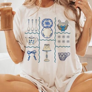 Pode incluir: Camiseta creme com um design de grade com ilustra&ccedil;&otilde;es azuis e brancas. O design inclui uma chaleira, pratos, colheres, uma galinha em uma cesta, uma l&acirc;mpada, uma x&iacute;cara de ch&aacute;, um la&ccedil;o e elementos florais. As ilustra&ccedil;&otilde;es t&ecirc;m uma est&eacute;tica vintage, desenhada &agrave; m&atilde;o.