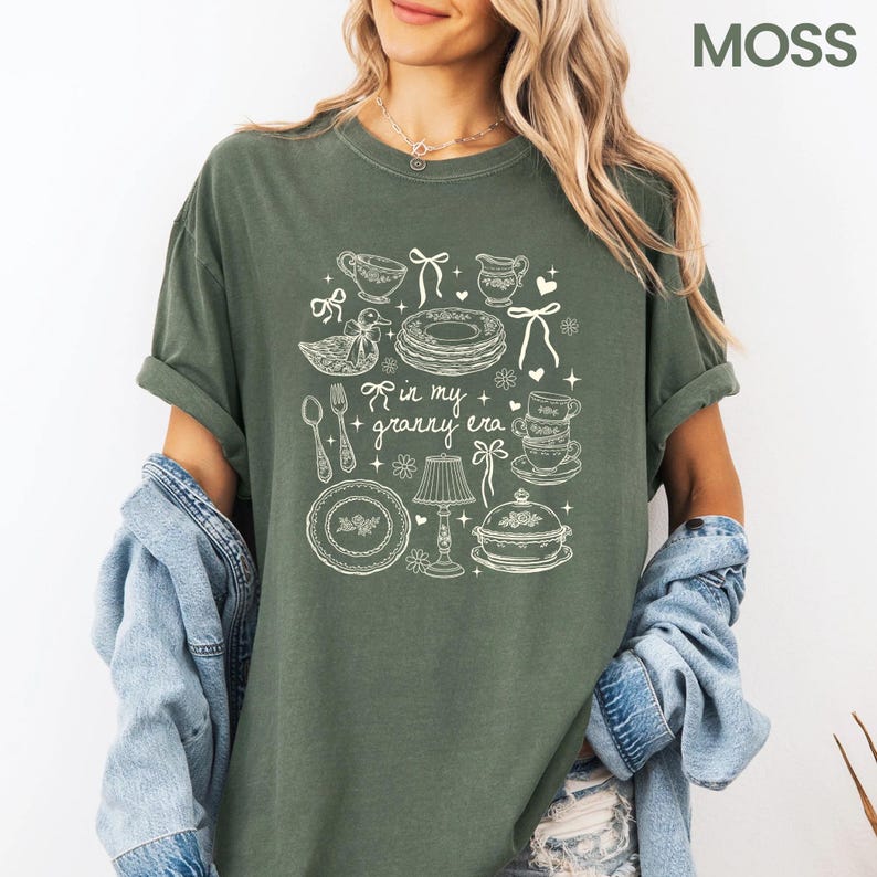 May include: A moss green t-shirt with a white graphic design featuring kitchenware illustrations, including plates, teacups, and a duck. The text "in my + granny era" is also included in the design.