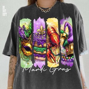 May include: Dark gray t-shirt featuring a colorful Mardi Gras design. The graphic includes a mask, cake, crawfish, and jester hat, with the words "Mardi Gras" in white script. The design is printed on a comfortable, casual tee.