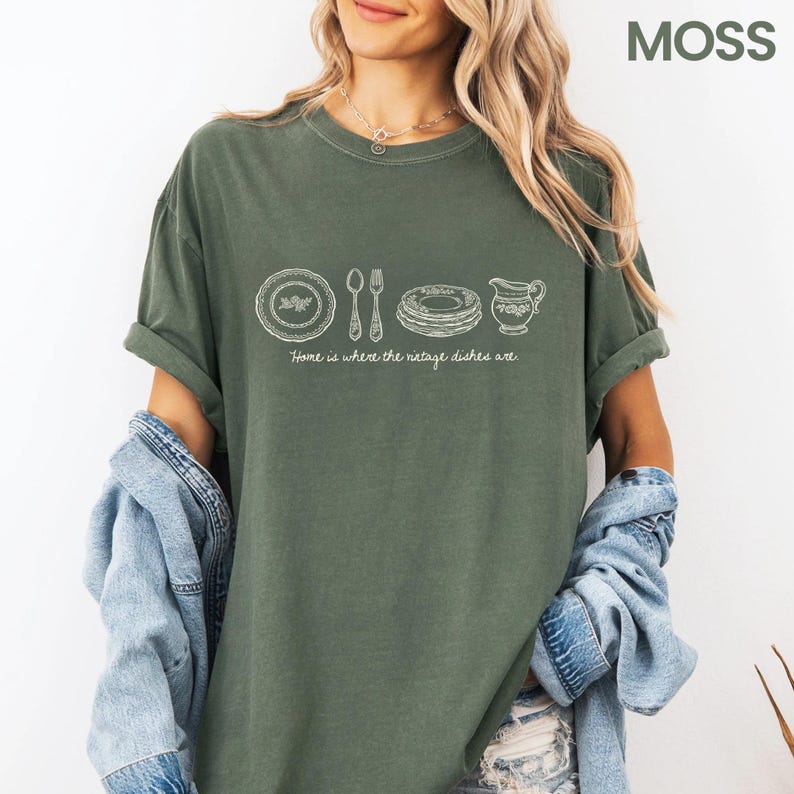 May include: Moss green t-shirt with a white graphic of vintage dishes, including plates, silverware, and a creamer. The text "Home is where the vintage dishes are" is below the graphic. The shirt has rolled-up sleeves and is paired with a denim jacket.