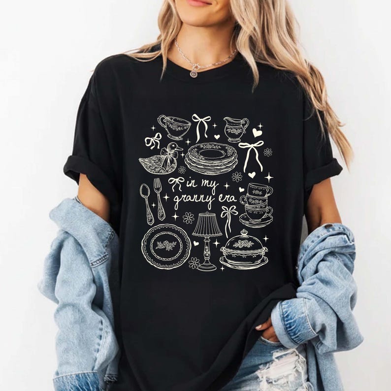 May include: Black t-shirt with a white graphic design featuring kitchenware illustrations, including plates, cups, and a lamp. The text "in my + granny era" is also included in the design. The shirt is a casual, comfortable style.