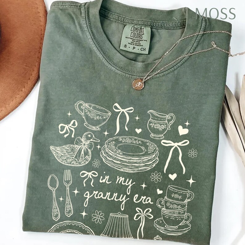 May include: A moss green Comfort Colors t-shirt with a white graphic design featuring teacups, plates, a duck, and the text "in my granny era." A silver necklace with a pendant is draped across the shirt.