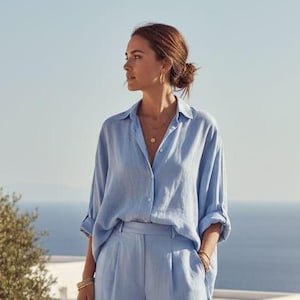 Könnte beinhalten: Ein hellblaues Leinen-Outfit, bestehend aus einem Hemd und einer passenden Hose mit weitem Bein. Das Hemd hat lange Ärmel und eine Knopfleiste. Die Hose hat eine hohe Taille und eine lockere Passform. Das Outfit wird im Freien mit Meerblick getragen.