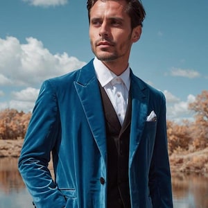 Puede incluir: Un blazer de terciopelo azul verdoso con chaleco negro y camisa blanca. El blazer tiene un cierre clásico de dos botones y un pañuelo de bolsillo. El hombre está de pie cerca de una masa de agua con cielo azul y nubes al fondo.