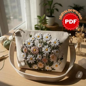 Op de afbeelding: Beige crossbodytas met een bloemenborduurontwerp met witte en gele madeliefjes en roze bloemen. De tas heeft een lange, verstelbare riem en goudkleurige hardware. De tas is gemaakt van canvas materiaal.