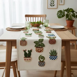Op de afbeelding: Een beige tafelloper met geborduurde potplanten in verschillende stijlen. De loper ligt op een houten tafel die gedekt is voor een maaltijd, met borden, glazen en een houten kom. De planten zijn in tinten groen, bruin en blauw.