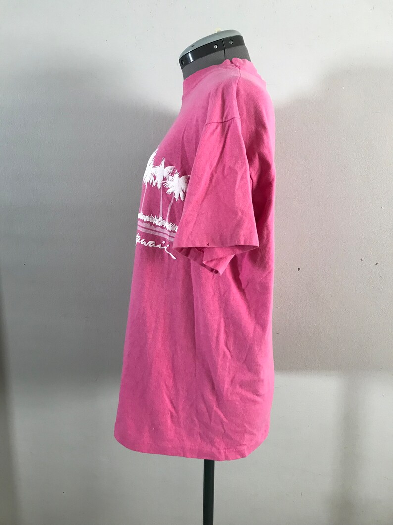 May include: Pink t-shirt with a white graphic of palm trees and the word "Hawaii" on the front.