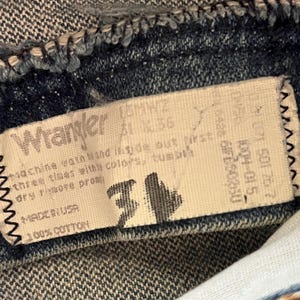 Puede incluir: Una etiqueta interior de una prenda de mezclilla marca Wrangler con el texto Wrangler, 100% cotton, Made in USA e instrucciones de cuidado. La etiqueta muestra la talla 31 en tinta negra grande, con c&oacute;digos y n&uacute;meros impresos en la tela blanca.