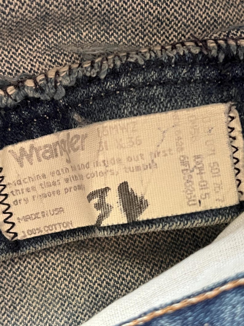 Puede incluir: Una etiqueta interior de una prenda de mezclilla marca Wrangler con el texto Wrangler, 100% cotton, Made in USA e instrucciones de cuidado. La etiqueta muestra la talla 31 en tinta negra grande, con c&oacute;digos y n&uacute;meros impresos en la tela blanca.
