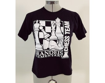 May include: Black t-shirt featuring a white graphic of chess pieces and a chessboard. The text "Brandeis Elementary Chess Team" is printed on the shirt.