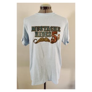 May include: Light blue t-shirt with the text "MUSTACHE RIDES 5¢" in a vintage style. The graphic includes a brown mustache and a red and brown "5¢" coin. The shirt is displayed on a mannequin.