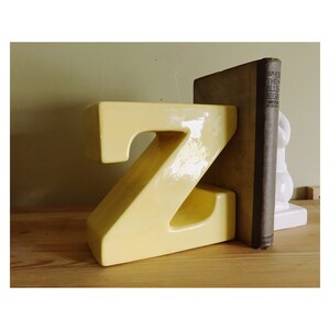May include: A yellow ceramic letter Z bookend. The bookend is sitting on a wooden surface and is holding up a brown book titled "When the Blue Begins".