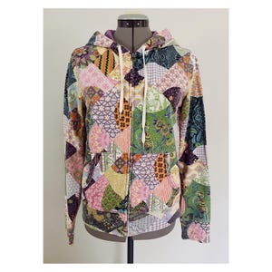 May include: A long-sleeve zip-up hoodie featuring a multi-colored patchwork design with various floral, geometric, and animal patterns in shades of green, purple, pink, orange, and black. The garment includes a hood with white drawstrings and two pockets.