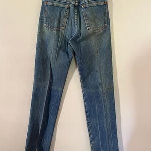 Puede incluir: Unos vaqueros de mezclilla azul con dise&ntilde;o de cintura alta, construcci&oacute;n cl&aacute;sica de cinco bolsillos, remaches de tono cobre en los bolsillos traseros, cierre de cremallera central y etiqueta de la marca Wrangler en el bolsillo trasero derecho.
