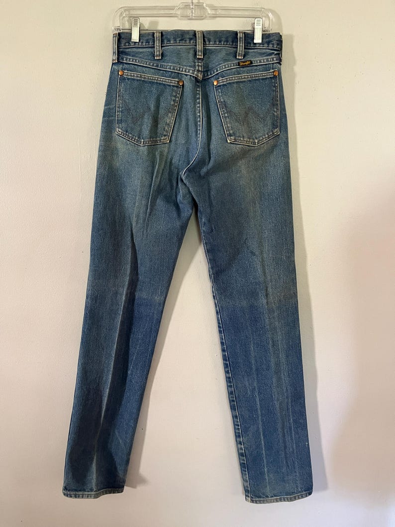 Puede incluir: Unos vaqueros de mezclilla azul con dise&ntilde;o de cintura alta, construcci&oacute;n cl&aacute;sica de cinco bolsillos, remaches de tono cobre en los bolsillos traseros, cierre de cremallera central y etiqueta de la marca Wrangler en el bolsillo trasero derecho.