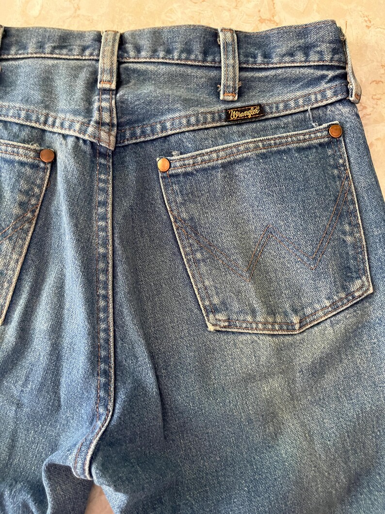 Puede incluir: Vaqueros de mezclilla azul con dise&ntilde;o de cintura alta, trabillas para cintur&oacute;n, canes&uacute; trasero y dos bolsillos traseros de parche. Cada bolsillo est&aacute; reforzado con remaches met&aacute;licos de color cobre y presenta una costura en forma de W. Etiqueta negra de Wrangler cosida.