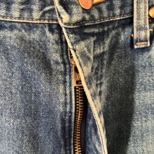 Puede incluir: Unos vaqueros de mezclilla azul con un acabado de lavado medio, una bragueta con cremallera met&aacute;lica y un bot&oacute;n de color cobre en la cintura grabado con el nombre de la marca Wrangler. La tela muestra una textura de sarga con costuras visibles.