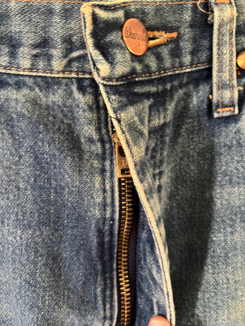 Puede incluir: Unos vaqueros de mezclilla azul con un acabado de lavado medio, una bragueta con cremallera met&aacute;lica y un bot&oacute;n de color cobre en la cintura grabado con el nombre de la marca Wrangler. La tela muestra una textura de sarga con costuras visibles.