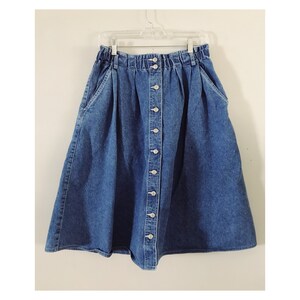 May include: A blue denim skirt with a button front and a gathered waist. The skirt has two pockets on the front and a slightly flared silhouette.