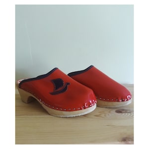 May include: A pair of red wooden clogs with a black bird design on the front. The clogs have a wooden heel and a leather upper.