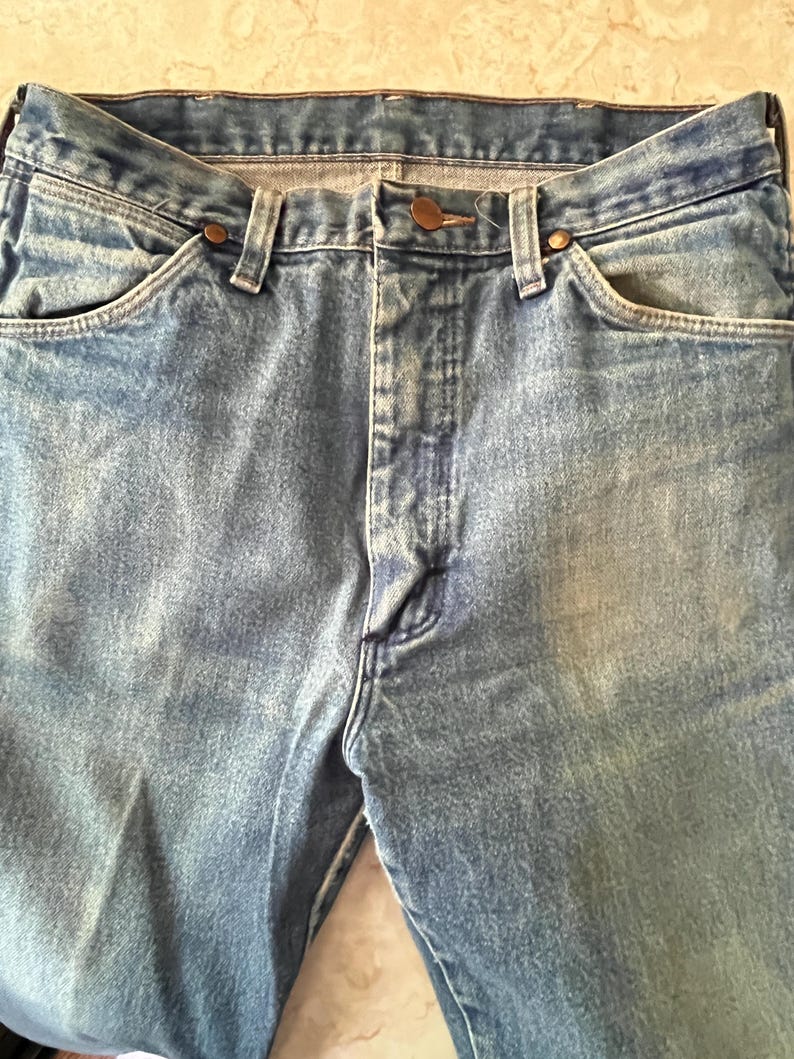 Puede incluir: Unos vaqueros de mezclilla azul claro con un dise&ntilde;o cl&aacute;sico de cinco bolsillos, cierre de botones y remaches met&aacute;licos de color cobre en las esquinas de los bolsillos. La tela presenta un lavado desgastado con costuras visibles y cintura.